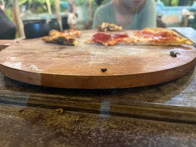 The image shows a close-up, low-angle view of a wooden pizza peel on a textured, dark brown table. Several slices of partially eaten pizza are scattered across the peel. In the foreground, on the table surface, there are small crumbs and a curved yellow object, possibly a piece of food. In the background, a person is partially visible, wearing a light green shirt, and other blurred figures are present in a setting that suggests an outdoor or semi-outdoor environment with greenery. The scene is consistent with a casual dining setting, possibly in Karimunjawa, Indonesia, given the tropical hints in the background.