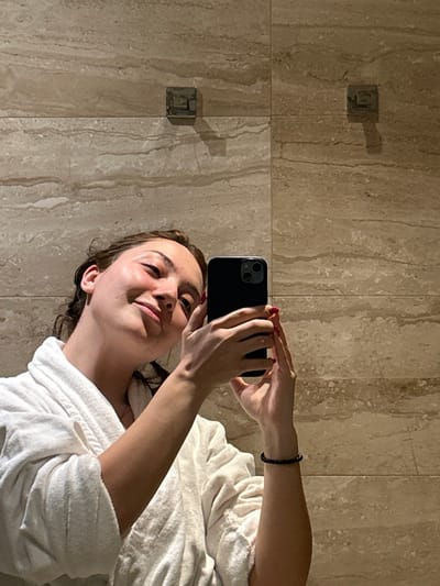 A woman in a white robe takes a selfie in a bathroom. She is holding a black smartphone, and her red-painted nails are visible. She has light skin and is smiling slightly. The bathroom features beige tiled walls, with shadows cast by the sunlight. Towel racks are visible on the wall, also casting shadows. The image is taken at an angle, making the scene appear sideways.