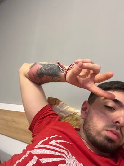 A forensic description of the scene in Erden, Bulgaria:

The image is a medium close-up, taken from a low angle, depicting a young adult male reclining indoors. The subject, positioned in the lower right and central frame, wears a red short-sleeved t-shirt with a white, claw-like graphic design partially visible on the chest. His left forearm, tattooed with a dark and red design (possibly a figure or helmet against a red backdrop), is raised. A red and white braided bracelet is worn on his left wrist. His left hand is presented with the thumb, index finger, and middle finger extended and spread, while the ring and pinky fingers are curled towards the palm. The individual has short dark hair and stubble, with his gaze directed forward, towards the camera. A patterned pillow, featuring light brown and beige tones, is partially visible behind his head. The background consists primarily of a plain light grey wall, with a light wooden panel or headboard visible on the far left. The lighting is consistent, suggesting an indoor artificial source.