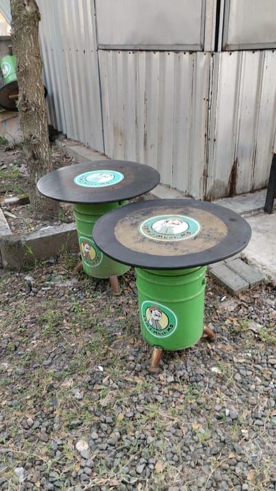 In Trosobo, Indonesia, two repurposed green oil drums serve as the bases for round tables. The table tops are black, circular discs, possibly made from vinyl records, with a sticker in the center featuring a cartoon character and text. The sticker displays a character wearing a hat and a shirt, with the words "STMJ Mbah Mo" around it. The tables are positioned outdoors on a ground covered with gravel, leaves, and sparse vegetation. Behind the tables is a corrugated metal wall, showing signs of weathering and rust streaks. A portion of a tree trunk is visible to the left of the tables. The scene appears to be an outdoor seating area or a roadside stall.