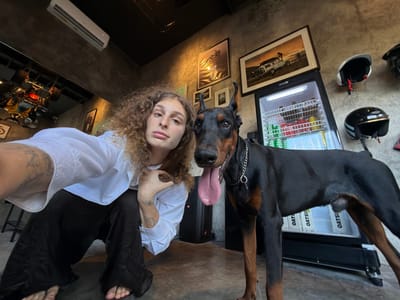 In the city of North Kuta, Indonesia, a close-up selfie captures a person and a Doberman Pinscher. The person, with curly brown hair and wearing a white collared shirt and black pants, is crouched on the left side of the frame, looking towards the camera with a neutral expression. Their left arm is raised, with their hand resting on their head, and tattoos are visible on their arm. The Doberman, standing to the right, faces the camera with its mouth open and tongue extended, wearing a silver chain collar. In the background, a black and tan cooler is filled with beverages. Above the cooler, framed pictures hang on a textured gray wall. To the left, a ceiling-mounted air conditioner unit is visible. The floor is a concrete surface.
