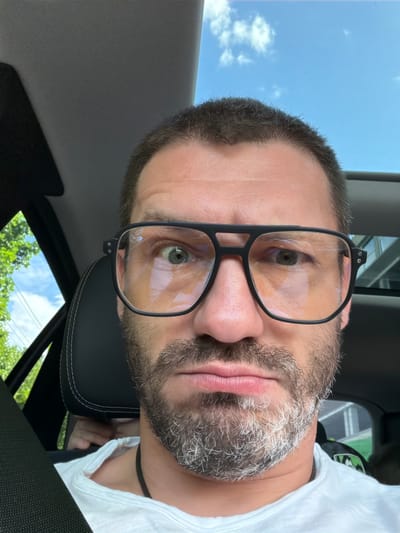 A close-up, selfie-style photo shows a man with short, dark hair, a beard with graying streaks, and large, black-rimmed glasses. He is wearing a white t-shirt. His left eye is wide open, and his right eye is slightly less so, with his eyebrows raised. His mouth is pursed. The man is sitting in the passenger seat of a car, as indicated by the car's interior visible in the background, including the headrest, a seatbelt, and a portion of the car's ceiling with a sunroof. Outside the car window, green trees and a blue sky with clouds are visible. The presence of a hand peeking out from behind the headrest suggests there may be another person in the backseat. The overall lighting suggests it is daytime. There is no discernible information in the image to indicate the location is in the city of Афанасово, Russia.