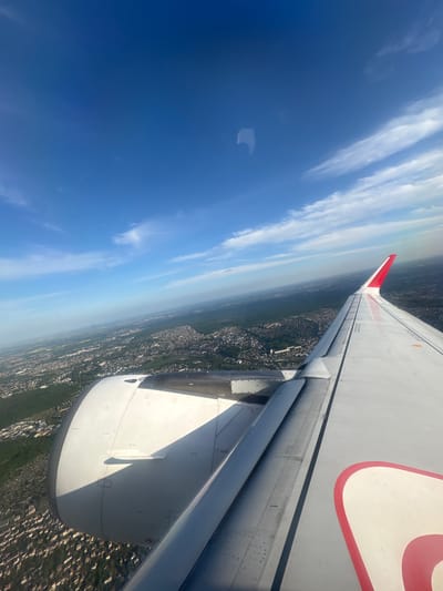 The view is from an airplane window showing the aircraft wing and engine with the city of Villeneuve-le-Roi, France below. The wing is predominantly white with a red accent. The engine is white and prominent in the foreground. Below, the landscape transitions from haze to discernible urban sprawl, indicating the ground level. The sky is blue with wisps of white clouds, suggestive of clear weather.