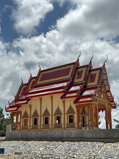 A Buddhist temple, likely under construction, is depicted in Thung Yai, Thailand, under a partly cloudy sky. The temple features a multi-tiered, vibrant roof with red, gold, green, and burgundy tiles, adorned with ornate golden finials that curve upwards. The facade is painted a pale yellow, accented with elaborate gold detailing around the windows and decorative pillars. Several windows with arched tops are visible, each framed by intricate golden patterns. A concrete retaining wall separates the temple from a lower area covered in grey stones. Two individuals are present near the temple, one appearing to be working. In the foreground, construction materials, including blue plastic sheeting and concrete forms, are scattered amongst the stones. Trees are visible on the left and right edges of the image.