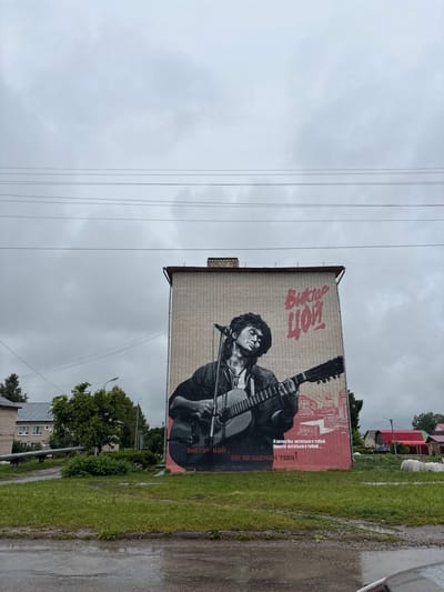 A mural of Viktor Tsoi, a Soviet rock musician, is painted on the side of a brick building in Sernur, Russia. Tsoi is depicted playing a guitar, with the name "Виктор Цой" (Viktor Tsoi) written in red above him. Below the portrait, a quote in Russian translates to "I wanted to stay with you, just stay with you... We will not forget you!". The building is located on a street with a grassy area in front, and residential buildings and trees are visible in the background. The sky is overcast and cloudy.