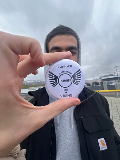 A young man is holding a white circular button in front of his face. The button has black text and imagery. The text on the button reads "Harbour Cafe, I ate fermented shark, I survived, I am a Viking". The man is wearing a grey hooded sweatshirt and a black Carhartt jacket. The man is standing in the town of Grundarfjörður, Iceland. The coordinates are 64.926 latitude and -23.256 longitude. The background is a cloudy sky and a paved area. The man is the only person visible in the photo.