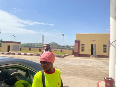 This is a vertical, rotated image taken in Pasere, Nigeria. The scene appears to be outdoors, possibly a street or open area adjacent to a building. The time of day is likely daytime, as indicated by the bright sky and clear lighting. The weather seems to be sunny with some scattered clouds.
In the foreground, a person is visible wearing a bright yellow shirt and a pink headscarf. They are looking towards the camera. Behind this person, another person can be seen, also looking towards the camera. They are positioned near a car.
The background features a building with yellow walls and dark red trim. This building has several windows and an open doorway. There is also a sign on the building with red text that appears to be in Arabic script. Further in the background, there are green fields and hills, suggesting a somewhat rural or semi-urban environment.
Other visible objects include a red bucket with the word "FIRE" written on it in white letters, attached to a white pole. There is also a lamppost visible.
The overall impression is a casual, everyday scene. The orientation of the image suggests it may have been taken with a phone held sideways.