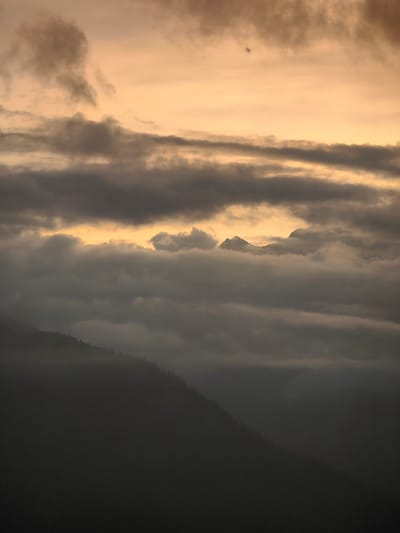 The image depicts a mountainous landscape with a dramatic sky. The foreground is dominated by a dark, silhouetted mountain slope covered in sparse trees, suggesting a steep incline. Above this, layers of thick, gray clouds obscure the view, with hints of lighter clouds and patches of golden light breaking through from behind. In the distance, the peaks of more mountains are visible, partially shrouded in mist and clouds, with a bright, hazy yellow light emanating from the horizon, indicating either sunrise or sunset. The overall atmosphere is moody and atmospheric, with a limited color palette of grays, blacks, and warm yellows. The specific location in the city of Aleo, India is not discernible from the visual elements present in the image.