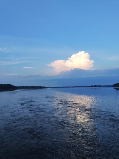 The image depicts a wide river under a vast, partly cloudy blue sky. The water is calm with a gentle ripple, reflecting the light of the sky and a large, bright cloud. Dark green trees line the distant banks of the river, creating a natural horizon. The scene appears to be set at either dawn or dusk, given the soft lighting and the color of the sky. There are no visible people or structures in the image, suggesting a natural, undeveloped landscape. The location in Edoma, Russia, is not discernible from the visual information presented.