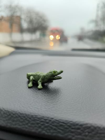 A small, green, plastic toy alligator, approximately 5-7 cm in length, with prominent yellow-green eyes and an open mouth displaying teeth, is positioned on a dark, textured dashboard in the immediate foreground. The background, viewed through a wet windshield, is heavily blurred, indicating precipitation. An outdoor urban or suburban road environment is discernible, with indistinct vehicles in the mid-distance, including one with two prominent, circular, yellow-orange headlights, possibly a truck or bus with a faint red upper body. Blurred outlines of buildings, including a light brown structure on the left, and bare trees line the roadside. The scene depicts an overcast or rainy daytime condition. The specific location within Illintsi, Ukraine, cannot be definitively identified from the visual information presented.