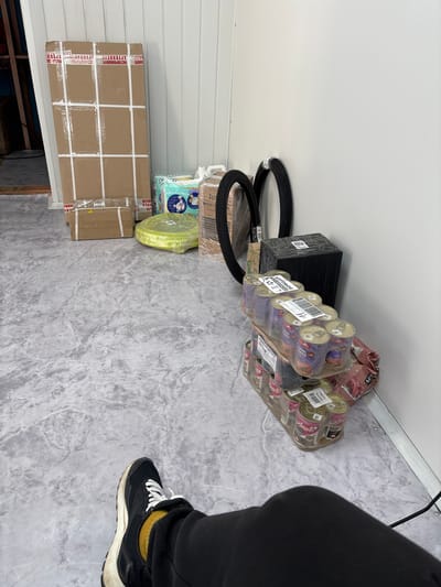 The image depicts a cluttered storage area, possibly a warehouse or a back room, with various items stacked and placed on a marble-patterned floor. In the foreground, a person's legs and feet are visible, wearing black pants and sneakers with white laces and a yellow sock. The person is seated, suggesting a moment of rest.
Dominating the background are several large cardboard boxes, some sealed with white tape and red warning labels. One particularly tall box is leaning against a white corrugated wall. To the right of the boxes are two bicycle tires, one partially wrapped in plastic.
In the mid-ground, a stack of canned goods, likely pet food based on the labels visible on some cans (e.g., "АППП"), is arranged in a plastic-wrapped pallet. Next to the cans is a black, rectangular object, possibly a speaker or storage unit, wrapped in plastic. A light green, coiled object, possibly a hose or cable, is also present. A package of diapers is visible between the large cardboard boxes and the tires.
The environment appears to be indoors, with a white corrugated wall forming the backdrop. The lighting is even, suggesting artificial illumination. The overall impression is one of storage or waiting for delivery/shipment. There is no direct indication of the city of Vileyka, Belarus within the image itself; this information would need to be provided externally to contextualize the scene.