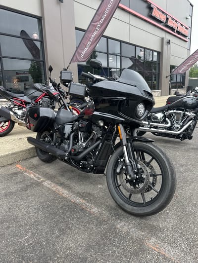 This media file captures a close-up, tilted view of several black motorcycles parked outside a Harley-Davidson dealership. The dealership's building is visible in the background, with large windows and a prominent sign reading "Barnes Harley-Davidson" in orange lettering. Two large banner flags with "BARNES HARLEY-DAVIDSON" printed on them are also present, one hanging from the building and another in the foreground.
The motorcycles are the primary subjects. The front of a large touring motorcycle is prominently featured, with its black fairing, headlight, and wheel. To its right, another motorcycle, a sportbike with red accents on its wheels, is partially visible. The motorcycles appear to be parked on an asphalt surface, with faint orange and white lines suggesting a parking area.
The scene appears to be outdoors during daylight hours, with overcast or bright sky reflected in the building's windows. The overall impression is one of a commercial setting, likely a dealership showcasing its inventory. There are no people visible in the image. The focus is on the machines themselves and the branding of the dealership.
