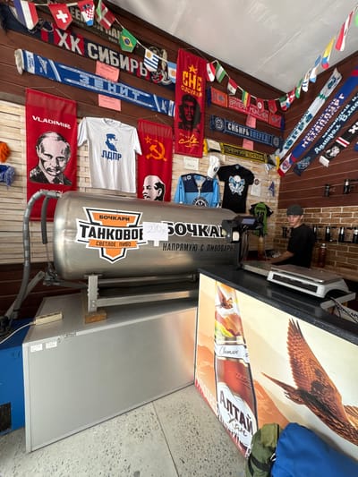 In the city of Ust-Sema, Russia, a scene unfolds inside a shop filled with merchandise, primarily sports-related memorabilia and clothing. Dominating the foreground is a large, silver cylindrical tank labeled "БОЧКАРИ ТАНКОВОЕ СВЕТЛОЕ ПИВО" (Bochkari Tank Light Beer), suggesting it is a beer dispenser. A price tag near the tank reads "210р". Behind the tank, a young male attendant wearing a dark t-shirt and a grey cap stands at a counter, looking towards the camera. The counter features a digital scale and several bottles of beverages. The walls and ceiling are adorned with numerous flags of various countries, sports team scarves, and posters featuring political figures like Vladimir Putin and Che Guevara, as well as athletic teams. T-shirts and jerseys are also displayed prominently. The environment appears to be a retail space, possibly a souvenir shop or a sports bar, with the overall impression of a bustling, eclectic establishment in Ust-Sema.
