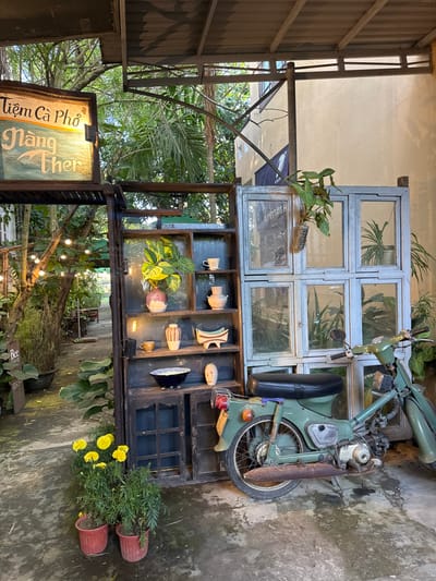 The image depicts an outdoor entrance area for a establishment named "Tiệm Cà Phố Nàng Ther" in Hội An Đông Ward, Vietnam. A dark wooden open shelving unit stands prominently on the left, displaying various ceramic items including vases, cups, and bowls, intermingled with potted green plants; several shelves are illuminated by small embedded lights. In the foreground, two terracotta pots contain yellow marigold plants. To the right of the shelving unit, a light-blue structure, constructed from repurposed window frames, serves as a display for additional potted plants behind its glass panes. A vintage-style, light-green moped with a black seat and visible rust on its exhaust pipe is parked in front of the blue structure. The ground is paved with worn, uneven concrete. In the background to the left, a path lined with trees and string lights recedes into a garden-like area. A light-colored building with a dark banner is partially visible in the right background. An overhead corrugated metal roof, supported by metal beams, provides partial cover for the scene.