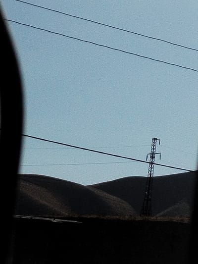 The image displays a desolate landscape under a clear sky, with rolling hills in the mid-ground. A solitary power line tower, constructed of lattice metal, stands prominently on the right side of the frame, its dark silhouette contrasting with the lighter sky. Multiple power lines stretch across the scene horizontally. The foreground is dominated by a dark, featureless expanse, possibly representing a road or the ground. The overall impression is one of rural isolation. The image does not contain any people or specific urban landmarks to indicate a precise location within the city of Kulob, Tajikistan, beyond it being a general outdoor scene that could be found in its vicinity.