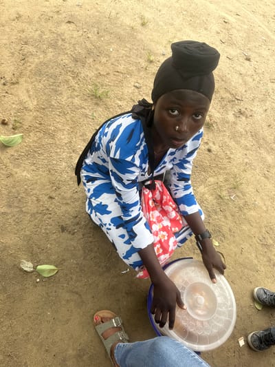 In Ganyingbo, Nigeria, a young person wearing a black headscarf and a blue and white patterned robe kneels on sandy ground. They are looking directly at the camera with a nose ring. To their right, a red bag with a white floral pattern is visible, along with a clear plastic container with a lid on the ground. Two pairs of shoes, one gray sneaker and one black sneaker, are partially visible on the ground to the right of the kneeling person. The ground is sandy with sparse vegetation.