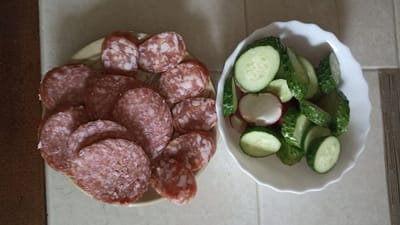 A top-down shot shows a light-colored surface with two plates of food. On the left, a round, shallow plate holds several slices of salami. The salami is reddish-pink with white fat marbling and appears to be thinly sliced. On the right, a white, ruffled bowl contains sliced cucumbers and radishes. The cucumbers are green with visible seeds and are cut into rounds and wedges. The radishes are white with a pinkish hue and are also sliced. The lighting is neutral, and the scene appears to be indoors. The image was taken in Kapyl, Belarus.