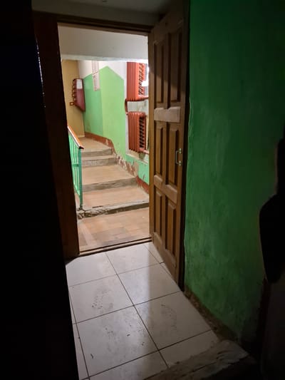 The image shows the interior of an apartment building hallway in Novlyanka, Russia. The hallway has a green-painted wall on the right and a wooden door open to the left. Beyond the open door, a staircase with a green railing is visible, leading upwards. The staircase and the floor in front of it are covered in terracotta tiles. There are radiators visible on the wall next to the staircase. The floor in the immediate foreground is tiled with white square tiles. A dark shadow obscures the lower left portion of the image.