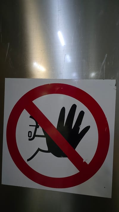 A "do not touch" sign is affixed to a metallic surface. The sign features a red circle with a diagonal line through it, superimposed over a black silhouette of a hand and what appears to be a numeral zero or letter 'o'. The metallic surface shows signs of wear, with scratches and reflections from overhead lighting. The image was taken in the city of Crailsheim, Germany.