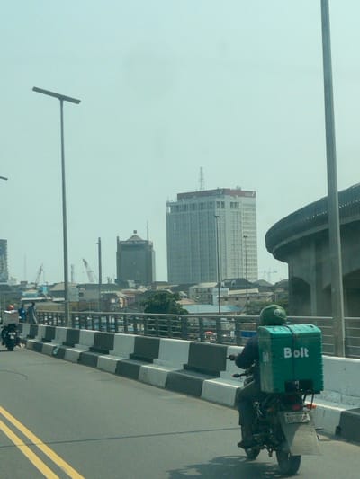 The image presents an urban street scene in Ebute-Metta, Lagos, Nigeria, under a hazy, bright sky. A paved road, marked with a double solid yellow line, occupies the lower portion of the frame. On the right, a motorcyclist wearing a green helmet and dark attire operates a motorcycle fitted with a large, rectangular, green delivery box prominently branded "Bolt" in white lettering. The motorcyclist is positioned with their back to the viewer, moving away. To the left of the road, a series of black and white concrete barriers separates the roadway from a narrow pedestrian area where several indistinct figures are visible. Further along the road on the left, another motorcyclist is discernible, also moving away, with a lighter-colored delivery box on their vehicle.
The background features a dense cityscape. A prominent tall, light-colored skyscraper with "UBA" branding near its roof stands in the center-right. To its left, a slightly shorter, darker multi-story building marked "WEMA BANK" is visible. Numerous other smaller buildings and some sparse vegetation populate the mid-ground. In the far distance on the left, at least two industrial cranes are visible, suggesting construction activity. A large, curving concrete overpass or ramp structure extends into the frame from the right, partially obscuring the horizon. Several tall, dark grey poles, likely streetlights, punctuate the scene vertically.