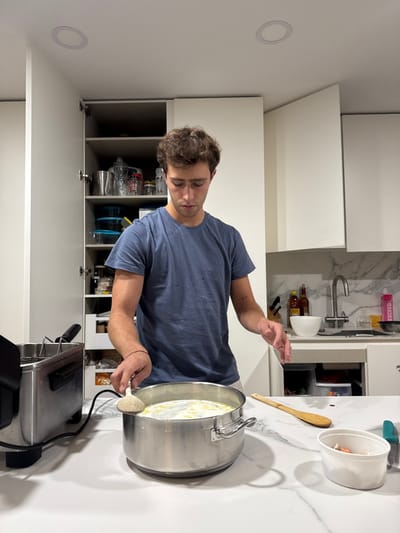 The scene is a modern kitchen in Cidade da Maia, Portugal, characterized by white flat-panel cabinetry, white marble-patterned countertops, and recessed ceiling lights. A male individual, appearing to be in his late teens or early twenties with brown curly hair, stands facing forward, looking down at a large silver metal pot situated on the countertop. He wears a blue t-shirt and light-colored shorts or trousers, with a red string bracelet on his left wrist and a small tattoo on his left forearm.
His left hand holds a wooden ladle submerged in the pot, which contains a light-colored liquid with white solid components. To the left of the pot, a silver deep fryer with a black power cord is partially visible. To the right, a long wooden spoon lies flat on the countertop, and a small white plastic container holds food scraps, positioned next to a knife with a teal handle. The background features open white wall cabinets revealing various kitchen items like containers and metal pitchers. A sink with a gooseneck faucet is visible on the right side of the background, alongside a white bowl, several bottles, and a pink cylindrical object labeled "EXCEED."