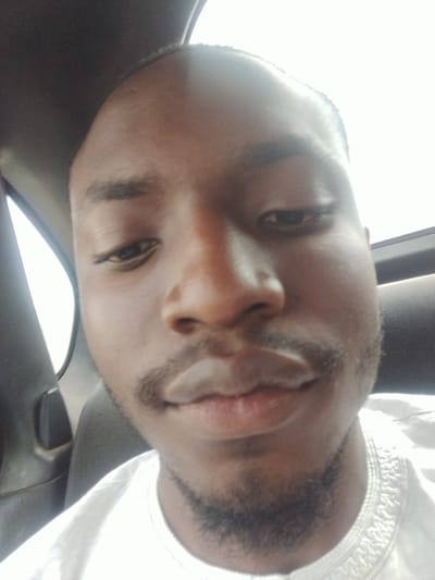 The media file is a close-up, selfie-style photograph of a young Black man's face, taken inside a vehicle. The man is looking directly at the camera, with his head tilted slightly upwards. He has dark skin, short hair, and a light mustache and goatee. His eyes are open and looking forward. He is wearing a white garment with an embroidered collar.
The lighting in the image is bright and somewhat overexposed, particularly on the right side of his face and forehead, suggesting it might be daytime. The background is blurred but shows the interior of a car, with a dark seat and a window providing light. The overall impression is that the photograph was taken casually, possibly while traveling. There is no visible text in the image. The location context provided is Jiru, Nigeria.