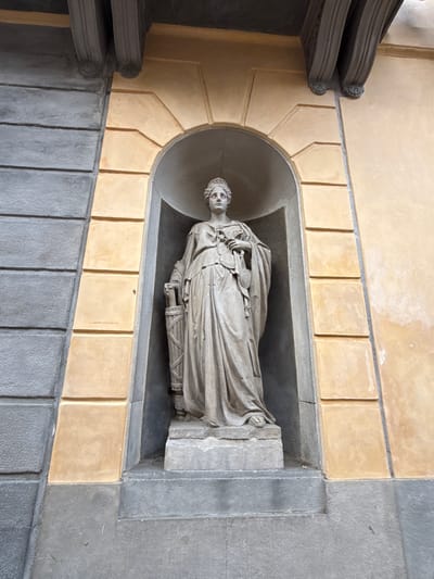 A stone statue of a robed female figure stands in an arched niche on a building facade in Mataró, Spain. The statue depicts a woman wearing a crown and holding scales and a scepter. She is dressed in a flowing gown. The niche is framed by textured walls, with a grey, horizontally segmented section on the left and a yellow, stucco-like section on the right. Above the niche, decorative architectural elements, possibly corbels, are visible. The base of the statue rests on a stepped stone pedestal. The scene appears to be an outdoor architectural feature of a historical building.