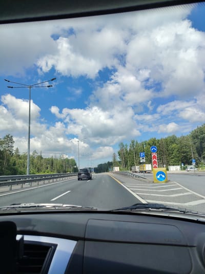The image depicts a view from inside a vehicle traveling on a highway. The sky is filled with scattered white clouds against a blue backdrop. Tall streetlights line the left side of the highway, which is bordered by a metal guardrail and a dense forest. A dark-colored SUV is visible ahead on the right lane. On the right side of the highway, there are several road signs, including parking symbols and directional arrows indicating a split in the road. Trucks are parked or driving in a separate lane on the right. The foreground shows the dashboard and windshield wipers of the vehicle. The scene appears to be in a rural or suburban area with extensive tree cover. This is likely the highway near Okulovka, Russia, given the general appearance of a Russian highway with roadside forests and typical road markings.