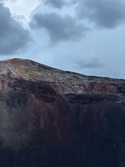 An image captures a volcanic landscape under a cloudy sky. The foreground is dominated by dark, rough volcanic rock, which transitions into a lighter, more varied slope of reddish and yellowish hues further up the mountain. A small, dark figure, potentially a person or an animal, is visible standing on the crest of the volcano. The sky is filled with heavy, gray clouds, suggesting overcast weather. There is no discernible urban environment or indication of the city of Unknown, Nicaragua within the frame.