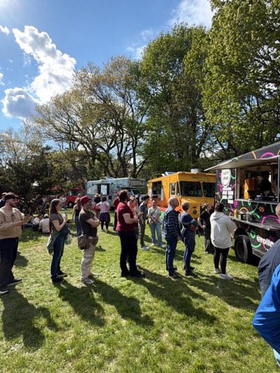The image depicts a scene in Rochester, United States, likely at a food truck rally or event. Several food trucks are parked in a grassy area, with a line of people waiting to order from one of them. The food trucks are various colors, including yellow and white, and feature different branding. A crowd of people is gathered in the area, some standing in line, others sitting on the grass. There are trees in the background, and the sky is partly cloudy. The atmosphere suggests a casual and festive gathering.