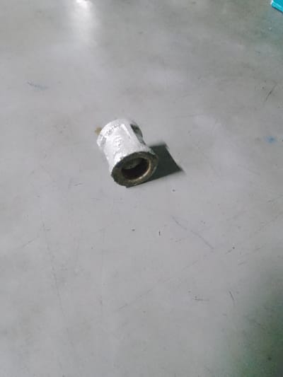 The image shows a single, metallic object, possibly a plumbing fitting, lying on a flat, gray surface. The object is cylindrical with openings at both ends and appears to be made of a shiny, textured material. It is positioned slightly off-center in the frame, casting a subtle shadow on the surface beneath it. The surface itself is plain and textured, with some faint lines and smudges visible. The lighting suggests an indoor setting, possibly with overhead light sources creating some bright reflections on the surface and object. There is no indication of people, animals, or any other discernible activity. The setting appears to be a functional space, perhaps a workshop, garage, or industrial area. No text is visible in the image. The scene is static and the overall mood is neutral, focusing solely on the solitary object.