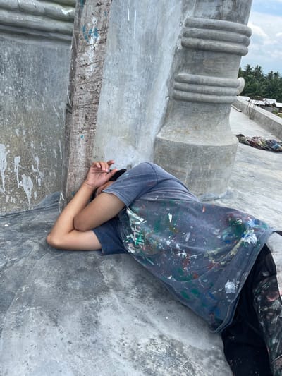 A male individual is observed lying on their left side on a grey concrete surface, leaning against a weathered concrete pillar in Kumbang, Indonesia. Their face is obscured by their right forearm and hand, which are bent and positioned over their head. The individual is wearing a dark blue t-shirt and dark pants, both extensively splattered with various colors of paint, including white, green, red, and blue. The concrete surface appears worn with minor cracking. To the right of the individual, a larger, barrel-shaped grey concrete architectural element with horizontal rings is visible. In the background, under a partly cloudy sky, a dense cluster of palm trees and distant low-rise structures are present. A piece of patterned fabric or tarp is also visible further along the concrete ground in the background. The scene is outdoors and appears to be daytime.