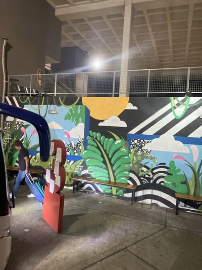 A man pulls a blue suitcase alongside a mural featuring vibrant flora, a yellow sun, and abstract geometric shapes. Nearby, a red structure with papers is visible. The background showcases a textured concrete wall and a ceiling with a grid-like pattern. A car with visible exhaust pipe is on the bottom of the frame. The image is taken at night with artificial lighting.