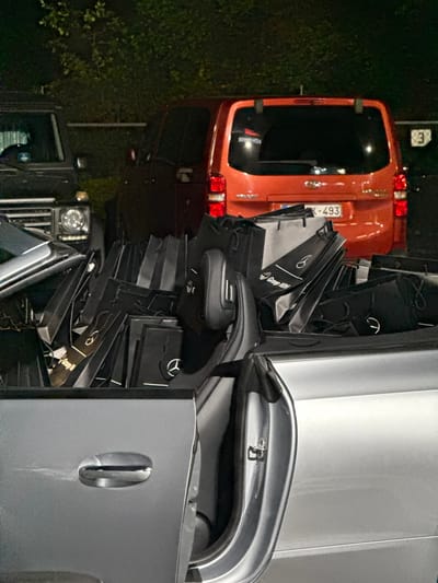 The scene depicts a parking area, likely an automotive dealership at Lierseweg 221, Herentals, Belgium, observed during nighttime or low-light conditions.

In the foreground, a silver (or light grey) convertible car is parked, with its passenger-side door ajar. The rear seating area and trunk space of this vehicle are completely filled with a large quantity of black shopping bags. Many of these bags feature the white Mercedes-Benz star logo, and at least one bag clearly displays "Group M. Van Mossel," a Mercedes-Benz dealership group in Belgium.

Behind the convertible, two other vehicles are parked. To the left, a dark-colored SUV, consistent with a Mercedes-Benz G-Class, is partially visible. To the right, an orange passenger van (possibly a Peugeot Partner/Rifter) is parked with its rear facing the viewer. This van exhibits a Belgian license plate, "1-K-493." A reflective sticker or text, possibly "BORN TO YOU," is visible on its rear window.

In the background, behind the parked vehicles, dark foliage is present, and a white sign with the number "3" is partially visible to the right of the orange van. No people are visible in the image.