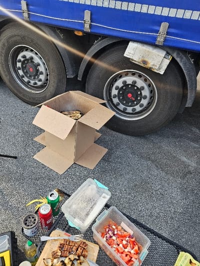 The image depicts an outdoor cooking and dining scene on dark grey asphalt pavement in Ventimiglia, Italy. The upper portion of the frame features the rear section of a commercial truck, characterized by a blue tarpaulin-covered trailer side secured by silver buckles, and two large black tires with silver hubcaps and red central caps. A black mudguard is positioned above the right tire, and a light-colored, folded fabric object secured above it displays burn marks on its lower edge. A bright sun flare diagonally crosses the upper left quadrant.
Centrally located is a makeshift grill fashioned from a brown cardboard box, containing visible glowing embers or charcoal and emitting smoke. Flat cardboard pieces form a base beneath this structure. The lower half of the image displays various items arranged on a black wire rack or directly on the ground. These include a wooden cutting board holding grilled, cubed meat (appearing to be chicken or pork) and some grilled onions, with a silver fork resting on it. Adjacent is a clear rectangular plastic container filled with chopped red tomatoes and white onion. Another larger, clear plastic container with a light blue lid is also visible. Two open aluminum beverage cans, one red and one green, are present. A small bottle of olive oil, a dark cylindrical object (possibly an ashtray or small burner), and a yellow tool are also discernible. A portion of a black object is visible on the far left. No individuals are present in the scene.