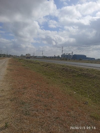 A rural scene features a road running horizontally across the midground. A truck travels along the road towards the left. Beyond the road are industrial buildings. Overhead power lines extend across the sky. A dirt road borders the grassy area in the foreground. The sky is filled with white clouds against a blue backdrop. The photo was taken on February 5, 2026, at 09:16.