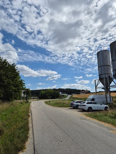 The image depicts a rural roadside scene in the vicinity of Mengkofen, Germany. A paved road curves through a landscape with tall green trees on the left, yellowing grass and wildflowers along the roadside, and fields beyond. In the background, a wooded hill rises, dotted with houses. Two large industrial silos stand prominently on the right side of the road, next to a white van and two parked cars, a silver Audi and a white Skoda. The sky above is a vibrant blue, filled with scattered white cumulus clouds. The overall impression is one of a quiet, sunny day in a rural setting.