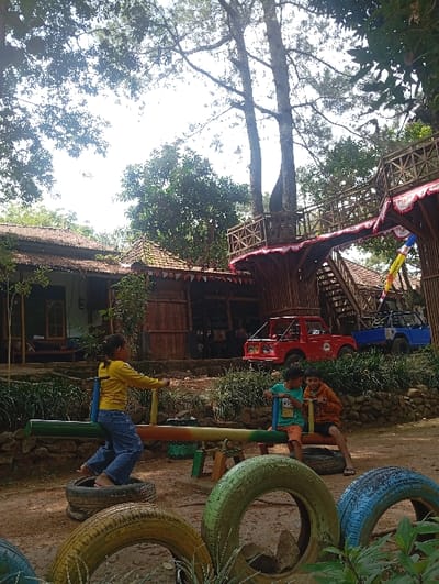 The image depicts an outdoor recreational area in Kajar, Indonesia, under bright daylight. Three children are positioned around a colorful seesaw on an unpaved dirt ground. On the left end of the seesaw, a barefoot girl wearing a yellow long-sleeved shirt and blue jeans is positioned to push down her side. On the right end, two boys are seated: one in a teal shirt and orange shorts, and the other in an orange long-sleeved shirt and dark shorts. The seesaw itself is painted with green, brown, and camouflage patterns, and its pivot points rest on used tires. Additional painted tires in yellow, green, and blue are visible on the ground in the foreground.

In the mid-ground and background, various structures are present. To the left, a traditional-style building with a dark tiled roof and wooden walls is visible. Further right, a larger, more elaborate elevated structure made of bamboo and wood features railings adorned with red and white banners. Two miniature off-road vehicles, one red and one blue, are parked on a raised dirt embankment behind the seesaw. The area is surrounded by dense green foliage, including large, tall trees with visible trunks and branches extending high into the bright sky.