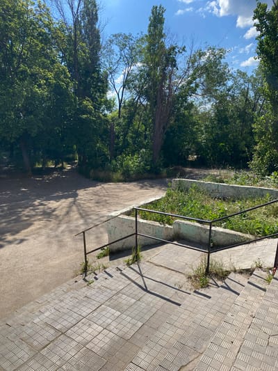The image depicts an outdoor scene on a sunny day, likely in a park or public area. In the foreground, a set of tiled stairs leads down from the viewpoint. A metal handrail with a dark finish is attached to a low, light-colored concrete wall that borders a raised planter bed filled with overgrown green vegetation. Beyond the planter, a paved path or walkway is visible, curving into a wooded area with dense green trees. Shadows from the trees are cast across the path, indicating bright sunlight. The sky above is blue with scattered white clouds. The overall impression is one of a somewhat unkempt but natural environment. The location is identified as Lopashchyne, Ukraine.