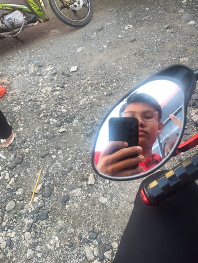 A close-up, high-angle shot captures a young man in glasses and a red shirt taking a selfie in a motorcycle's side-view mirror. He is holding a black smartphone, with his left hand visible. Another person's reflection is partially visible in the mirror. The motorcycle's handlebar, grip, and a red accent light are visible in the lower right corner. The ground is covered in gravel and small stones, with a hint of a motorcycle's engine and wheel in the upper left. A yellow stick lies on the ground in the center. The scene appears to be outdoors in a rural or semi-urban area, potentially in Pompanua, Indonesia, given the typical environment.