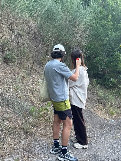 The image shows two individuals, one wearing a grey t-shirt and lime green shorts, and the other wearing a cream-colored oversized shirt and dark brown pants. The person in the t-shirt has a canvas tote bag slung over their shoulder and is wearing a white baseball cap. The person in the cream shirt has a red object, possibly a flower, being placed behind their ear by the other individual. They are standing on a gravel path next to a grassy, overgrown embankment. The environment appears to be a natural, rural setting, likely in Čučuci, Montenegro, characterized by dry vegetation and dense green foliage in the background. The lighting suggests it is daytime.