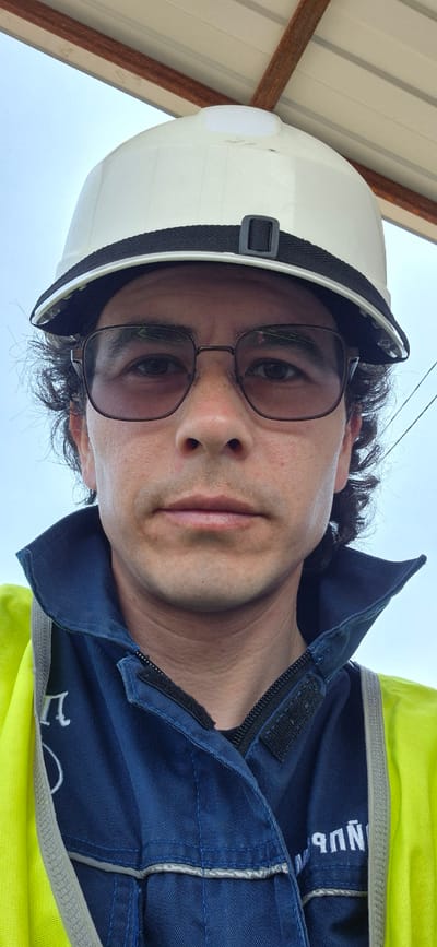 A man wearing a white hard hat and glasses is facing the camera, likely in Анастасьевка, Russia. He is wearing a blue work uniform with reflective accents and a yellow underlayer. Overhead, there is a metal structure, potentially a roof. Power lines are visible in the background. The overall perspective indicates the photo was taken from a low angle, looking up.