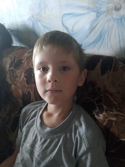 A young boy with blonde hair and blue eyes is sitting on a couch. He is wearing a gray t-shirt. The couch has a brown floral pattern. The background is a light-colored wall with a blue floral design. The image appears to be taken indoors. The location is identified as Изыхские Копи, Russia, which is a village in the Khakassia region.