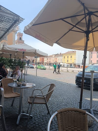 An outdoor cafe in Casalmaggiore, Italy, is depicted in the image. The foreground features a round metal table with two wicker chairs. On the table, there is a wine glass, a small bowl with some items, and an ashtray. In the background, a cobblestone street is visible, with several people walking. A green car is parked on the street, and buildings with colorful facades line the street. Two large umbrellas provide shade for the cafe seating area. The sky is clear and blue. A bell tower is visible in the distance, part of a building in the historic center of Casalmaggiore.