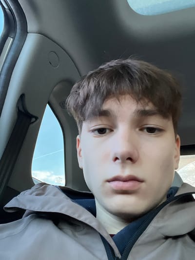A young male individual, with brown hair and light skin, is depicted in a front-facing, close-up shot, seated within the passenger compartment of a vehicle. He is wearing a light tan or brown collared jacket over a dark blue or navy garment, with a visible maroon layer beneath. His eyes are directed towards the camera, and his facial expression is neutral. To the left of the individual, the light grey interior trim of the vehicle's A-pillar is visible, featuring a black seatbelt strap and a small circular indent. Beyond the vehicle's window on the left, a clear blue sky is visible, traversed by a faint white contrail. The exact location within Zainsk, Russia, cannot be determined from the visual information presented in the image.