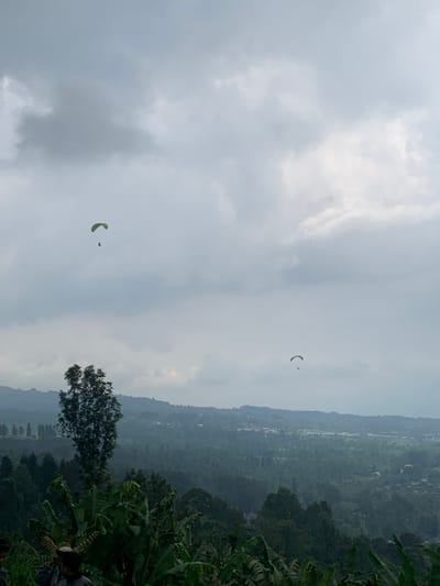 The image displays a panoramic view of a mountainous landscape in Puncak, Indonesia, under an overcast sky with varying cloud densities. Two paragliders are actively aloft; one with a light-toned canopy and a dark, suspended figure is positioned in the upper-left quadrant, while a second, smaller paraglider with a similar appearance is visible lower and to the right. The mid-ground and background feature multiple layers of rolling hills and mountains densely covered in dark green, tropical vegetation. A distant valley or plain below the mountains contains a scattered settlement with indistinct buildings and infrastructure. The immediate foreground consists of lush, green foliage, prominently featuring large, broad leaves consistent with banana plants, and the trunk of a tall, slender tree. Partially visible human figures are present at the bottom-left edge of the frame.