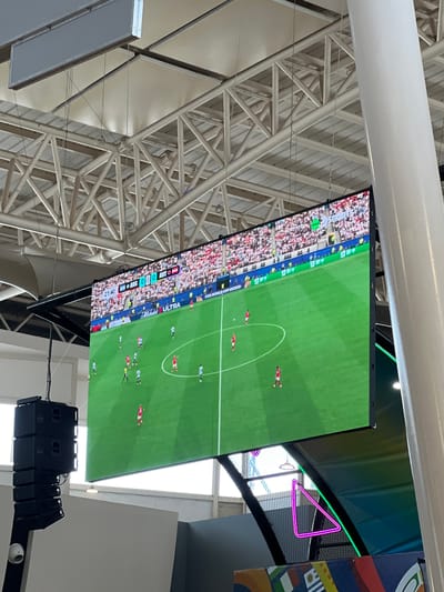 The media file shows a large screen displaying a soccer match. The screen is mounted high up, suggesting it's in a stadium, arena, or large indoor venue with exposed ceiling structures and white beams. A portion of the stadium seating, filled with a crowd of people wearing various colored clothing, is visible on the left side of the screen. The soccer field is green with white markings, and several players in red and light blue uniforms are scattered across it. The game appears to be in progress, with some players in motion.
At the bottom left of the screen, there's a score overlay. It shows "ARG 0" and "AUT 0," indicating the score is tied at zero. There's also a timer displaying "21:40." Text like "FIFA WORLD CUP" and "ULTRA" are visible on the screen.
To the right of the screen, a neon pink triangular light fixture is visible against a dark background, with a green light strip above it. The overall atmosphere seems to be that of a sports viewing event, likely indoors, given the lighting and ceiling structure. The time of day is not explicitly stated, but the indoor lighting suggests it could be anytime. There are no obvious weather cues. The presence of the crowd and the displayed game implies a public gathering for entertainment.