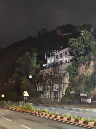 The image depicts a nighttime scene of a multi-story building constructed on a steep hillside. The building, which appears to be white, has several windows illuminated with warm, yellow light, suggesting occupancy. The hillside is covered with vegetation and rocky outcrops. At the base of the hillside, a road is visible with traffic markings. Road signs, including a triangular warning sign and a directional sign, are present along the roadside. The overall environment appears to be an urban or semi-urban setting, possibly in a hilly region. The lighting suggests it is dark, with artificial lights from the building and streetlights illuminating the scene.