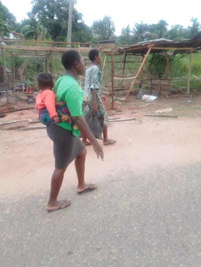 In Alayi, Nigeria, the image shows two adult women walking on a paved road, with a child secured to the back of the first woman with a patterned cloth wrap. The first woman wears a green t-shirt and a dark skirt, and the child is wearing a long-sleeved orange shirt. Both adults wear sandals. Debris and wooden structures are present in the background.