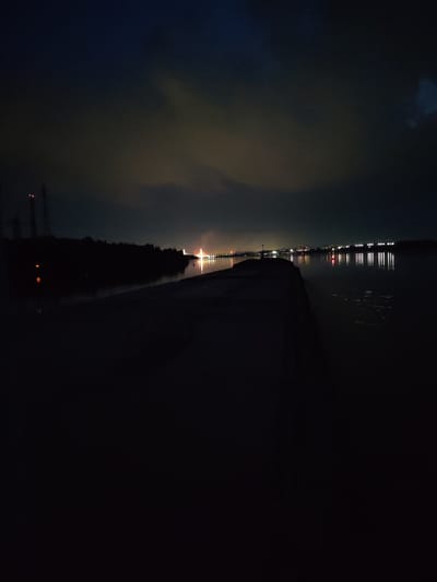 The image depicts a nighttime scene of a body of water with a dark, silhouetted structure in the foreground, possibly a pier or a breakwater. In the distance, across the water, a city skyline is visible, illuminated by artificial lights. The lights of the city reflect on the calm surface of the water. The sky above is dark with some diffused clouds, possibly illuminated by the city lights or distant lightning. There are no visible people or specific landmarks that would allow for the identification of the location as Lapach, Russia, or any other specific city. The overall atmosphere is dark and quiet.
